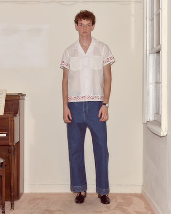 A man stands by a piano indoors, wearing wide blue jeans and the Cherry Lane Embroidered Shirt—a white cotton poplin men's shirt featuring red cherry trim accents.