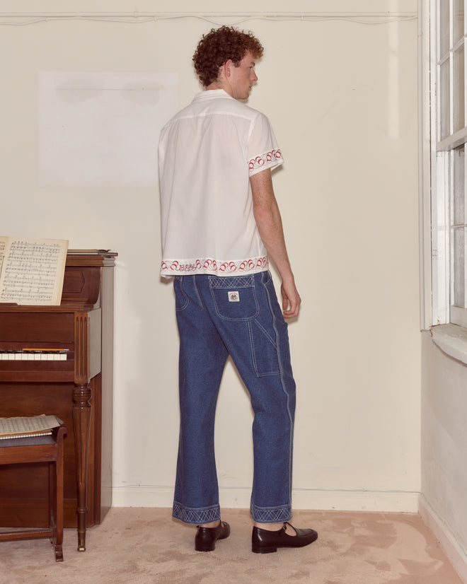 A man stands indoors by a piano, wearing blue jeans, black shoes, and the Cherry Lane Embroidered Shirt.