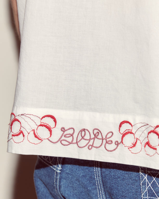 The Cherry Lane Embroidered Shirt is a white cotton poplin men's shirt with red cherries and "BODE" embroidered on the hem, styled here with blue jeans.