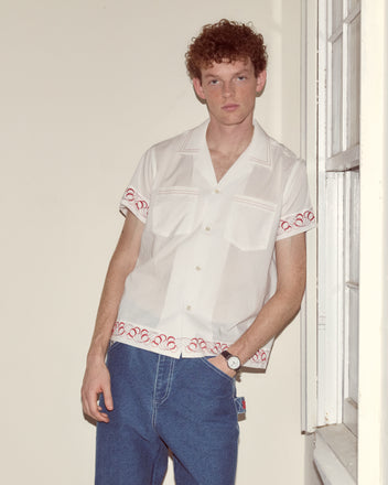 Wearing the Cherry Lane Embroidered Shirt and blue jeans, a man stands by an indoor window.