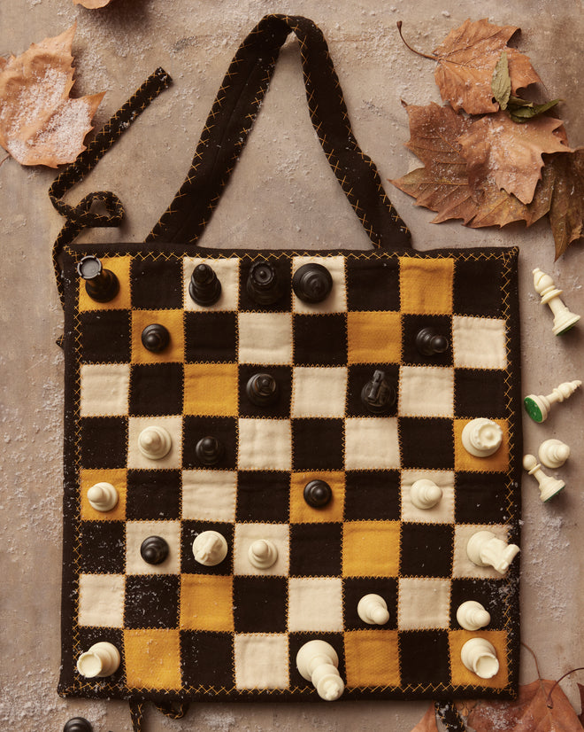 A Board Game Travel Set, featuring a quilted chessboard that brings to mind a patchwork quilt, is adorned with elegant chess pieces and surrounded by scattered autumn leaves.