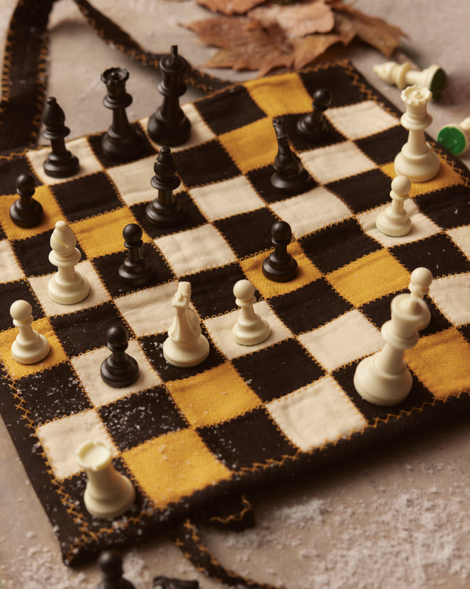 A Board Game Travel Set, featuring a quilted chessboard that brings to mind a patchwork quilt, is adorned with elegant chess pieces and surrounded by scattered autumn leaves.