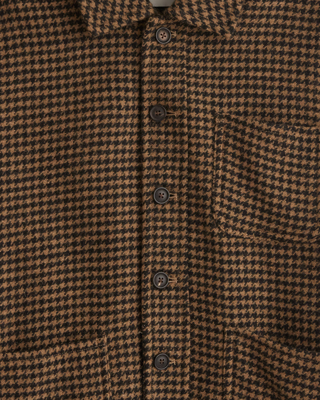 The Chestnut Hound Jacket - S/M, a brown plaid button-up crafted from 100% wool, is displayed on a hanger against a tiled wall, exuding a unique charm reminiscent of the elegance found in 1960s suiting fabrics.