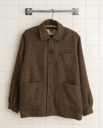 The Chestnut Hound Jacket - S/M, a brown plaid button-up crafted from 100% wool, is displayed on a hanger against a tiled wall, exuding a unique charm reminiscent of the elegance found in 1960s suiting fabrics.