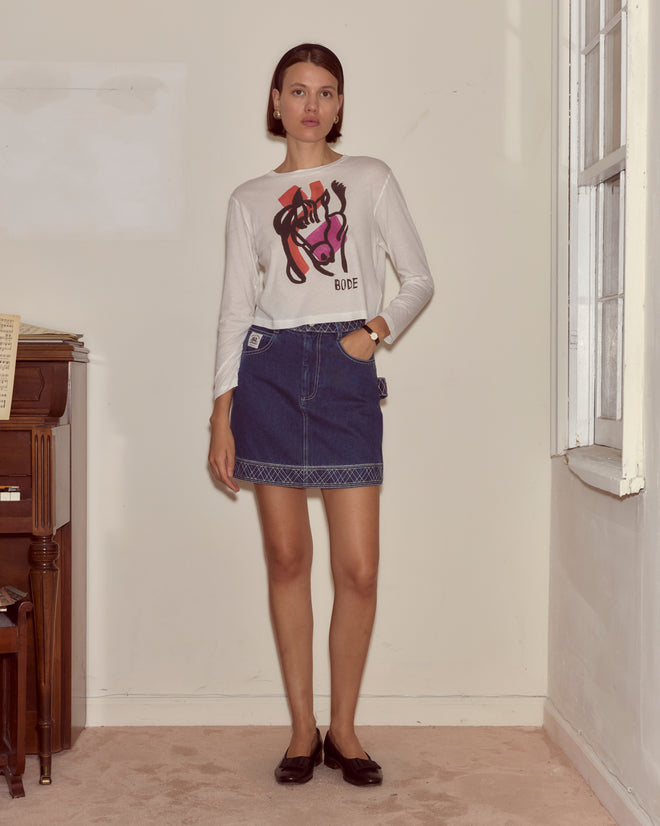 Woman wears the Cheval Long Sleeve Tee and a denim skirt while standing indoors by a piano and window, looking at the camera.