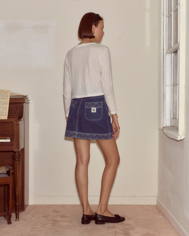 A woman stands indoors facing away, wearing the Cheval Long Sleeve Tee—white with abstract colorblocking—paired with a denim skirt and black loafers.