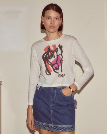 A woman stands against a beige wall wearing the Cheval Long Sleeve Tee with an illustrated horse motif and a blue denim skirt.