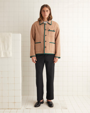 A person stands against a tiled background, wearing the Chevron Grid Jacket, a beige checked wool jacket with green trim, along with black pants and black shoes. A white towel hangs on a partial wall to the left, enhancing the workwear silhouette.