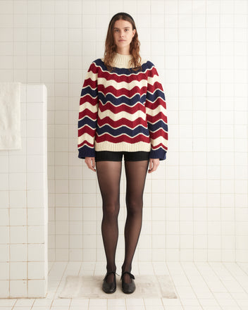 A woman stands in a tiled room wearing a Chevron Pullover with red, white, and blue chevron stripes, black shorts, black tights, and black shoes.