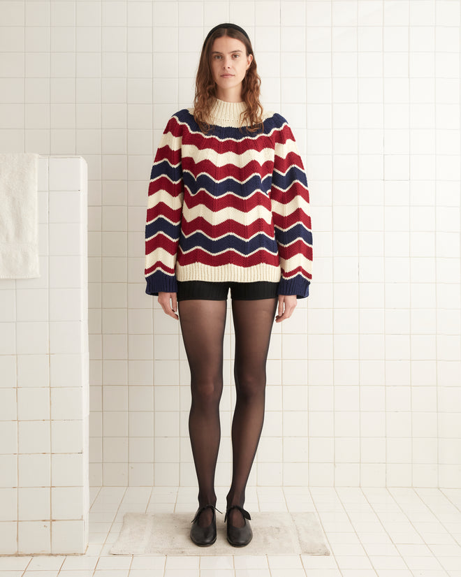 A woman stands in a tiled room wearing a Chevron Pullover with red, white, and blue chevron stripes, black shorts, black tights, and black shoes.