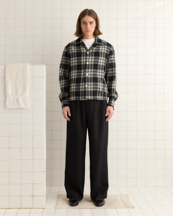 A person in a Chimney Plaid Overshirt - L/XL and black pants stands in a tiled room next to a towel.