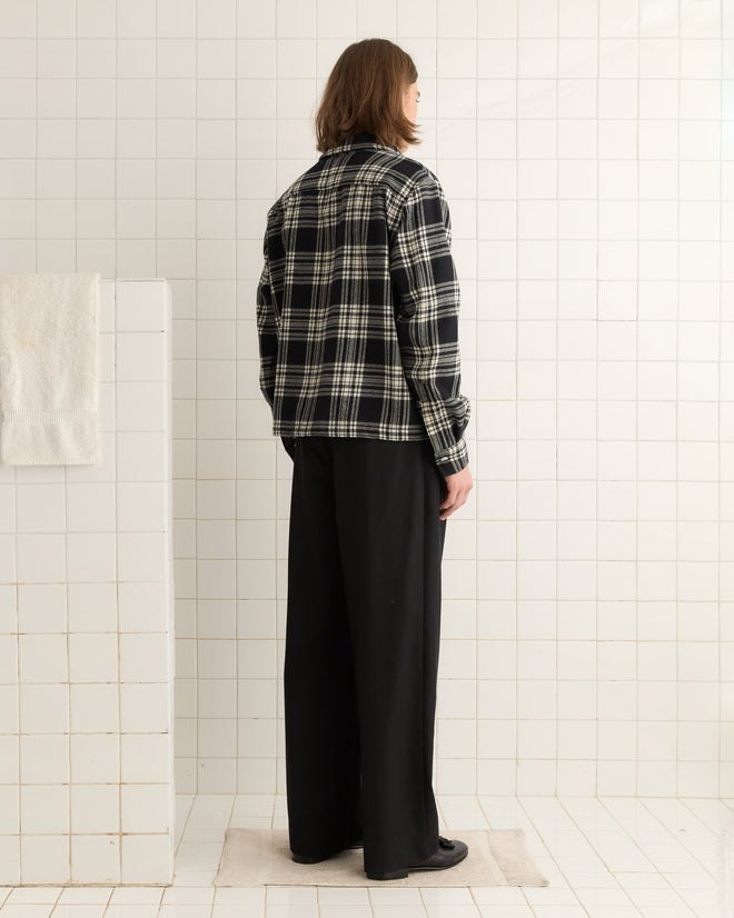 Person with shoulder-length hair wears the Chimney Plaid Overshirt - M/L in black and matching black pants while standing in a tiled room.