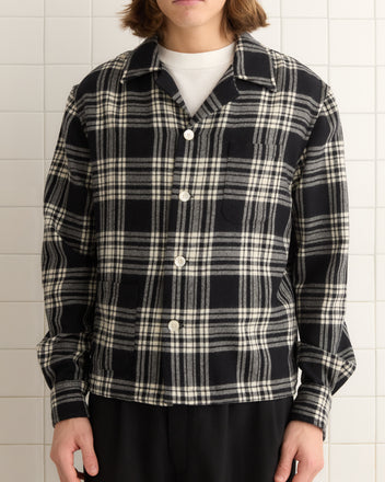 A person stands stylishly against a tiled wall wearing the Chimney Plaid Overshirt - L/XL, a unique black and white plaid button-up shirt over a white top.