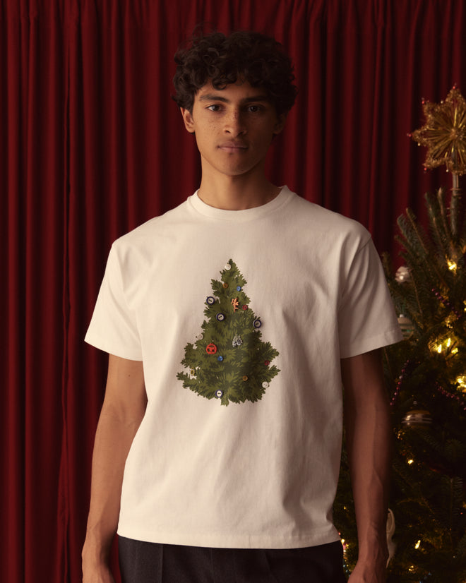 A person wearing the Christmas Tree Tee stands near a decorated tree, showcasing holiday styles against a rich red curtain backdrop.