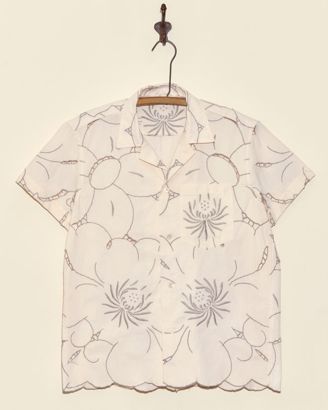 Chrysanthemum Carpet Short Sleeve Shirt (XS) in cream, featuring a bold, abstract floral pattern and button-up front, hangs on a hook.