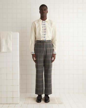 A person wearing the Cingoli Trousers - 32, styled with a white shirt featuring printed details, stands in a tile-lined room, highlighting unique textile craftsmanship and side buckle waist adjusters.