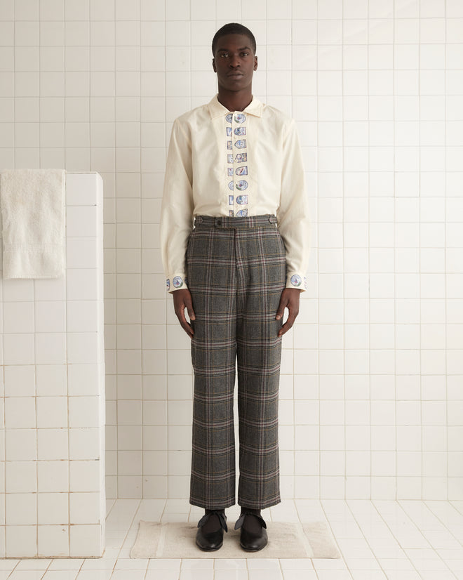 A person wearing the Cingoli Trousers - 32, styled with a white shirt featuring printed details, stands in a tile-lined room, highlighting unique textile craftsmanship and side buckle waist adjusters.