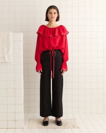 A person stands in a bathroom wearing a Satin Claret Blouse - Red with black pants, against a tiled wall.