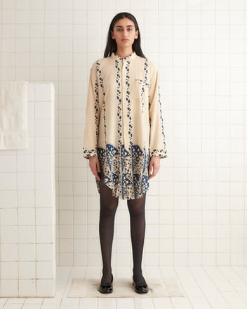 A woman in the Climbing Ivy Tunic, featuring a 1920s-1930s fabric design, stands on a tiled floor, facing forward against a tiled wall.