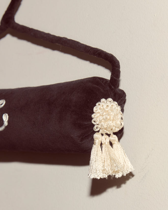 The Cloakroom Hanger - Black, featuring a black velvet cushion with white tassels, is suspended by an antique hanger against a light-colored wall.