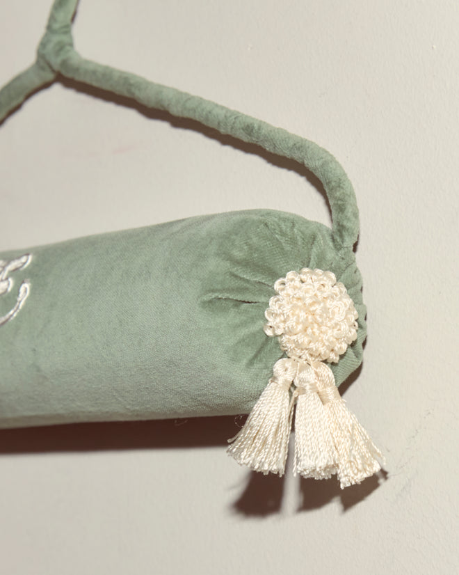 A green velvet bolster pillow with white tassel and rosette detail hangs from a Cloakroom Hanger - Teal, displayed against a plain wall.