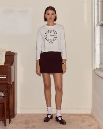 A woman stands indoors wearing a Clock Long Sleeve Tee made from vintage-weight cotton, paired with a black skirt and black shoes.