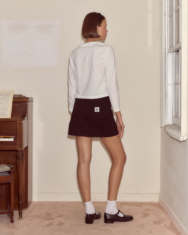 A woman in a white Clock Long Sleeve Tee and black skirt stands indoors, facing away near a window and piano.