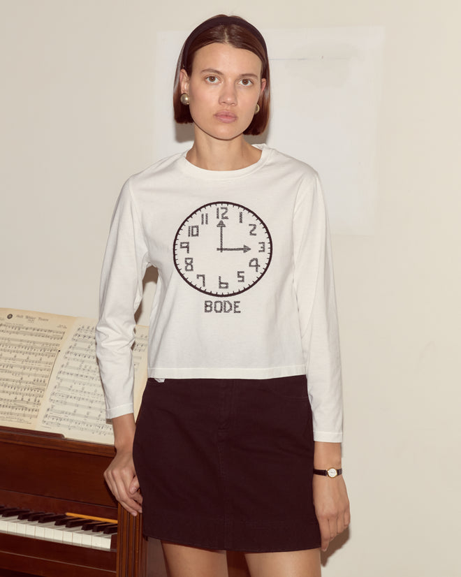 A woman wearing the Clock Long Sleeve Tee stands by a piano with sheet music, looking at the camera.