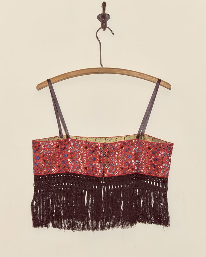 The Cloister Garden Bra in size S, featuring black antique piano shawl fringe, is displayed on a wooden hanger against a plain wall.