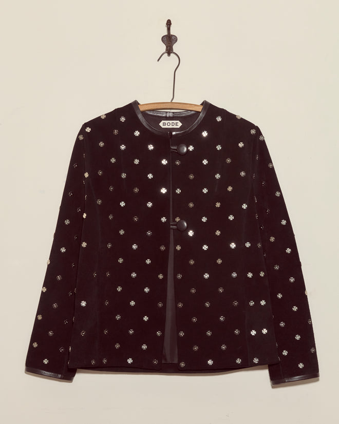 The Clover Charm Jacket, a black suede piece with small silver floral patterns and a boxy silhouette, hangs on a wooden hanger against a beige wall.