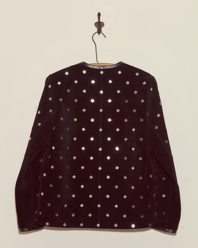 The Clover Charm Jacket, a vintage suede piece with shiny silver floral studs and a boxy silhouette, hangs on a hook against a plain light-colored wall.