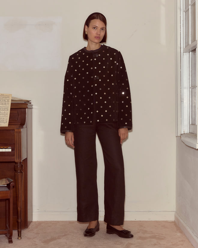 A woman wears the Clover Charm Jacket—a boxy black suede piece with small white dots—paired with black pants as she stands indoors by a piano and a window.