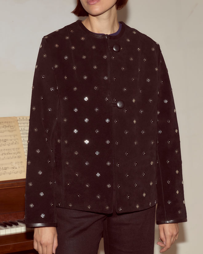 Person wearing the Clover Charm Jacket, a black suede piece with large buttons and a boxy silhouette, stands in front of a piano with sheet music.