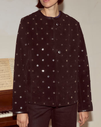 Person wearing the Clover Charm Jacket, a black suede piece with large buttons and a boxy silhouette, stands in front of a piano with sheet music.