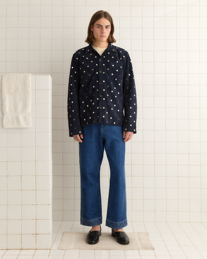 A person in a Clover Work Shirt - Dark Navy, echoing a midcentury style, and jeans stands in a tiled room with towels hanging on the side.