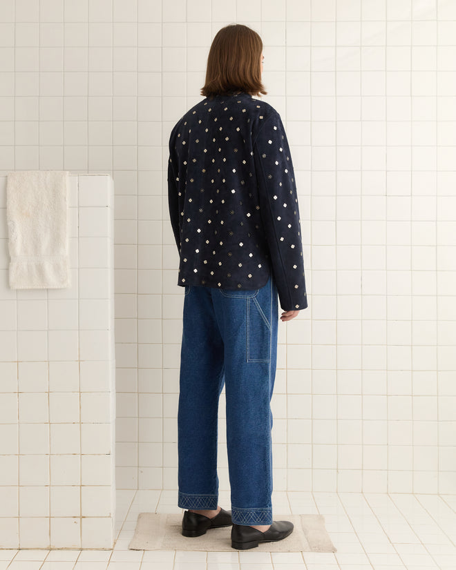 A long-haired individual in a polka dot jacket and jeans, accessorized with silver Clover Work Shirt charms, stands in a tiled room where a towel hangs on the wall.