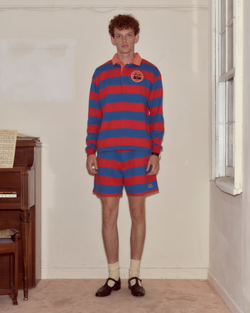 A man wearing the Clubhouse Rugby Shirt, featuring wide red and blue stripes, paired with shorts, socks, and black shoes stands indoors by a piano.