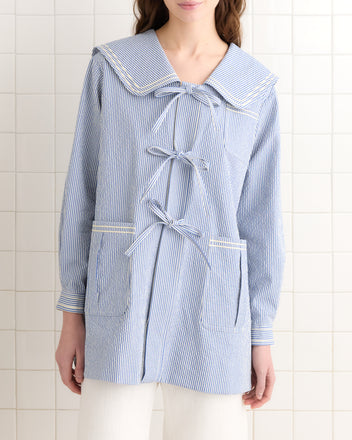 A woman wears the Club Stripe Sailor Coat in blue and white, featuring a large collar and three bows, standing against a tiled wall.