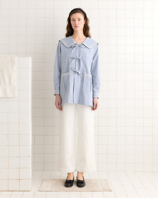 Woman wearing the Club Stripe Sailor Coat in blue and white, paired with white pants and black shoes, standing in a tiled room.