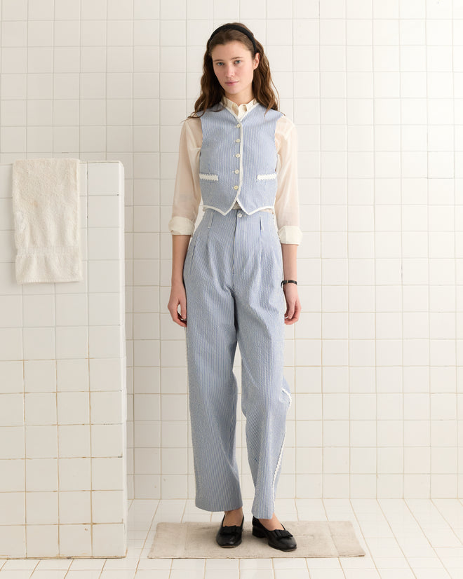 A woman wearing the Club Stripe Waistcoat in Blue/White and high-waisted pants stands in a tiled room with a towel hanging on the wall.
