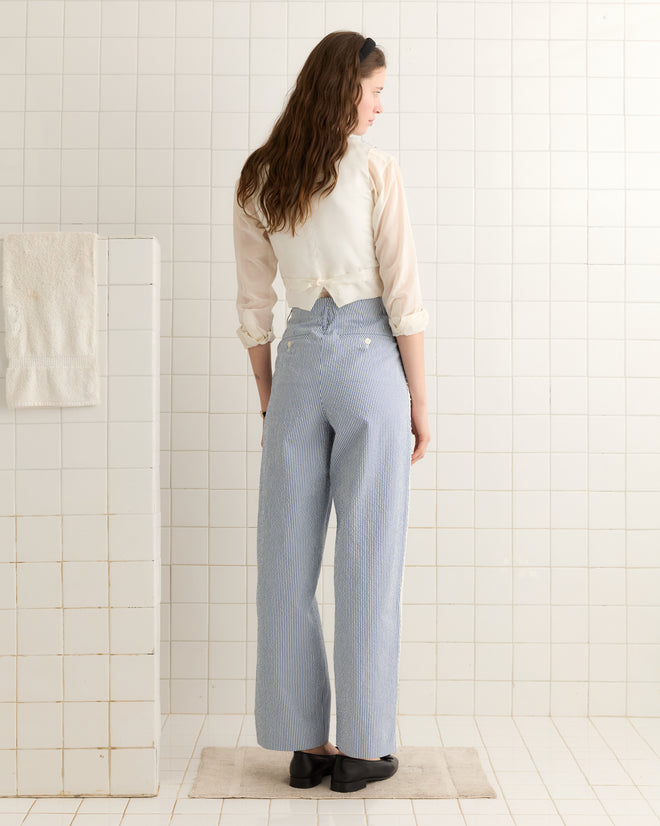 A woman with long hair faces a white tiled wall, wearing the Club Stripe Waistcoat in blue/white.