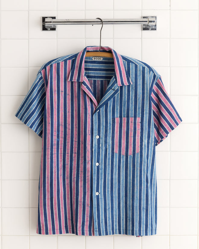 The Cobalt Coral Stripe Shirt - L/XL features vertical pink and blue stripes, crafted from unique deadstock fabric, shown hanging on a hanger against a tiled wall.