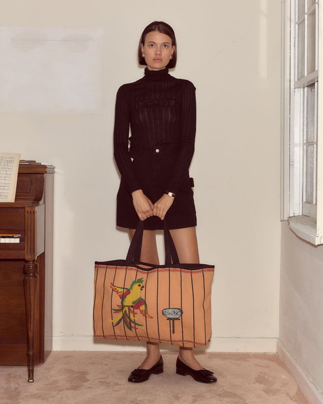 A woman in a black outfit stands indoors holding the Cockatoo Large Tote, a spacious tan canvas bag featuring a vibrant parrot design.