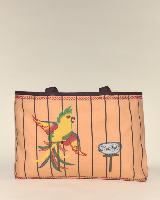 The Cockatoo Large Tote features tan canvas with vertical black stripes, a vibrant embroidered parrot, and a blue patterned accent—a colorful everyday tote.