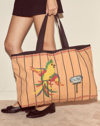 A person carries the Cockatoo Large Tote, featuring a colorful parrot and black stripes, styled with a skirt and black flats.