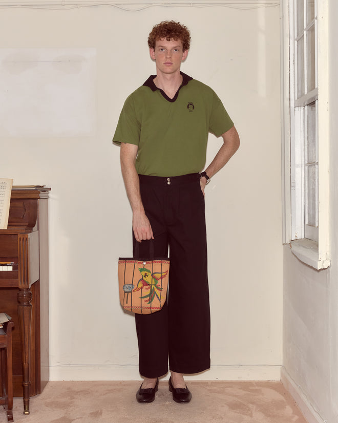 A person in a green shirt and black pants stands indoors by a piano, holding the Cockatoo Small Tote.