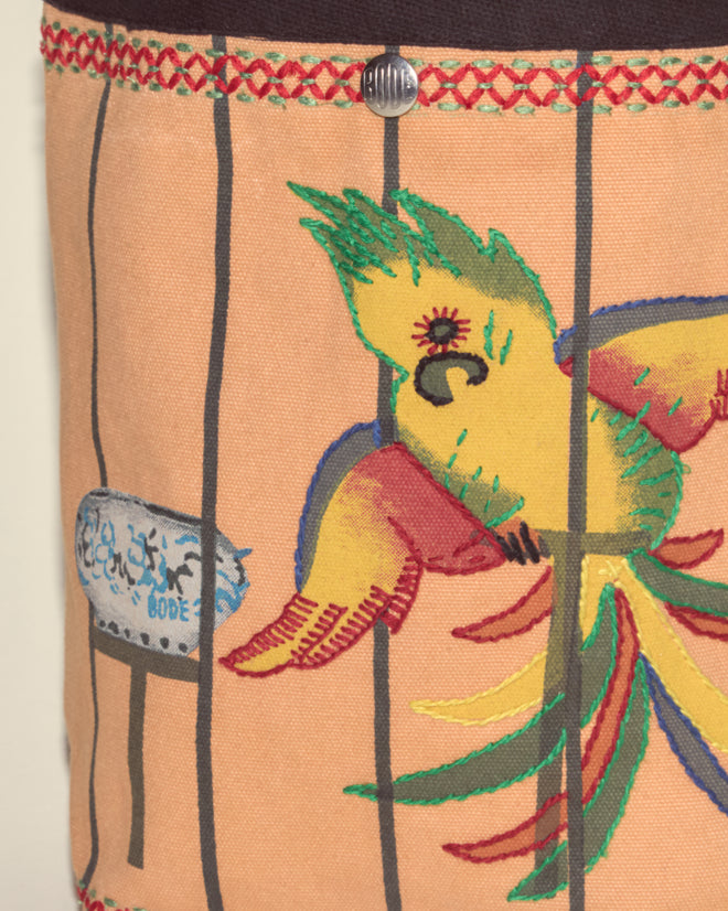 A colorful cockatoo is perched behind cage bars with a sign in the background, standing on patterned fabric resembling the Cockatoo Small Tote, which features a button closure at the top.