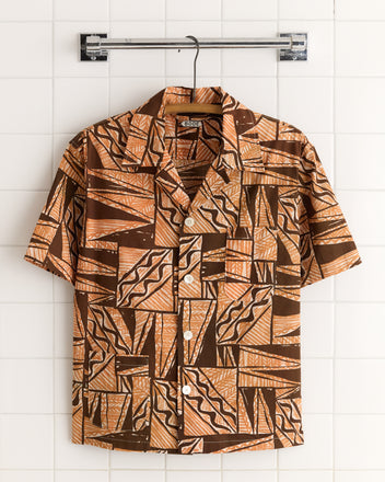 The Cocoa Bean Shirt - S/M is a short-sleeved button-up with a brown and tan geometric pattern and textured weave, displayed on a metal hanger.