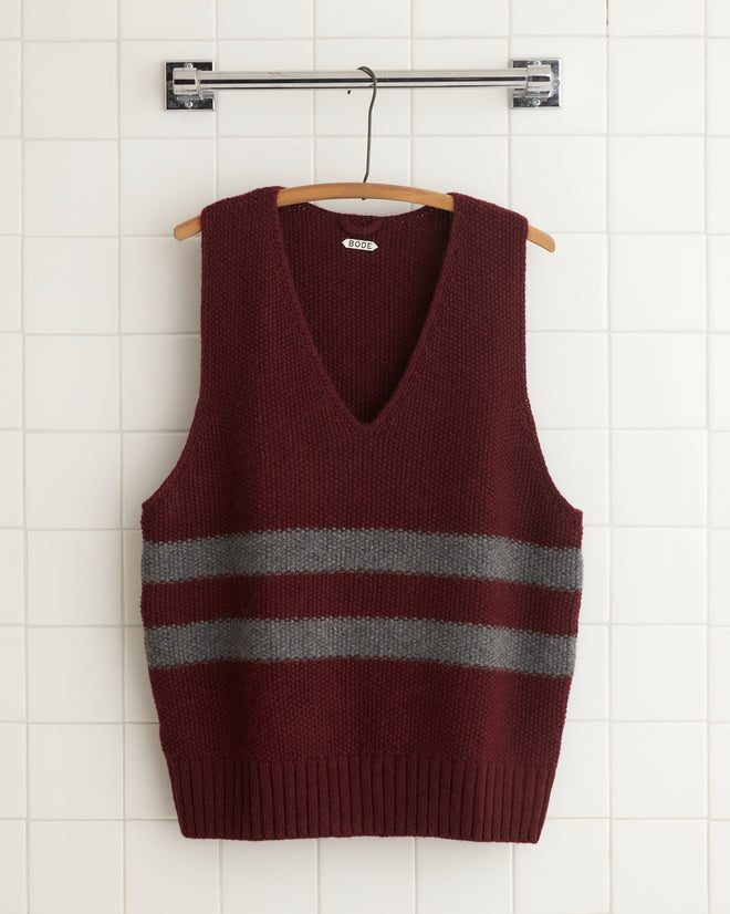 The Collegiate Vest, a maroon v-neck sweater vest crafted from soft alpaca wool, is accented with two gray stripes and displayed on a wooden hanger against a tiled background.