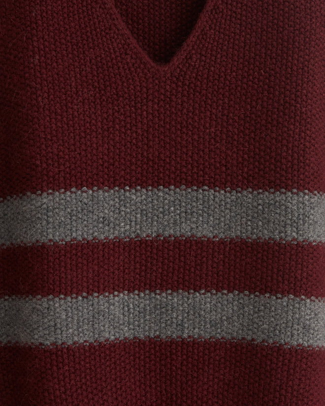 The Collegiate Vest, a maroon v-neck sweater vest crafted from soft alpaca wool, is accented with two gray stripes and displayed on a wooden hanger against a tiled background.